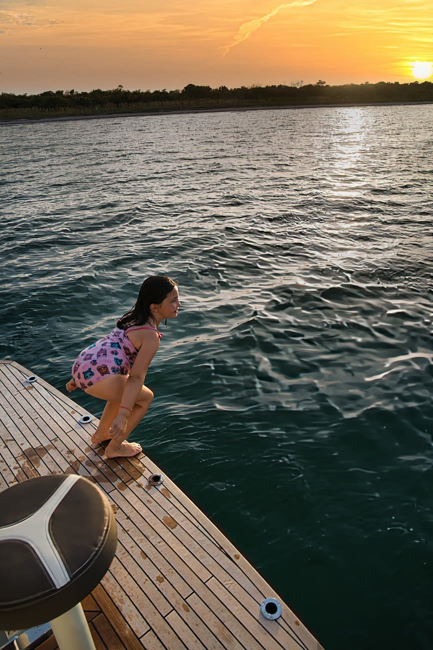 A child crouched on the teak swim platform at sunset, about to jump